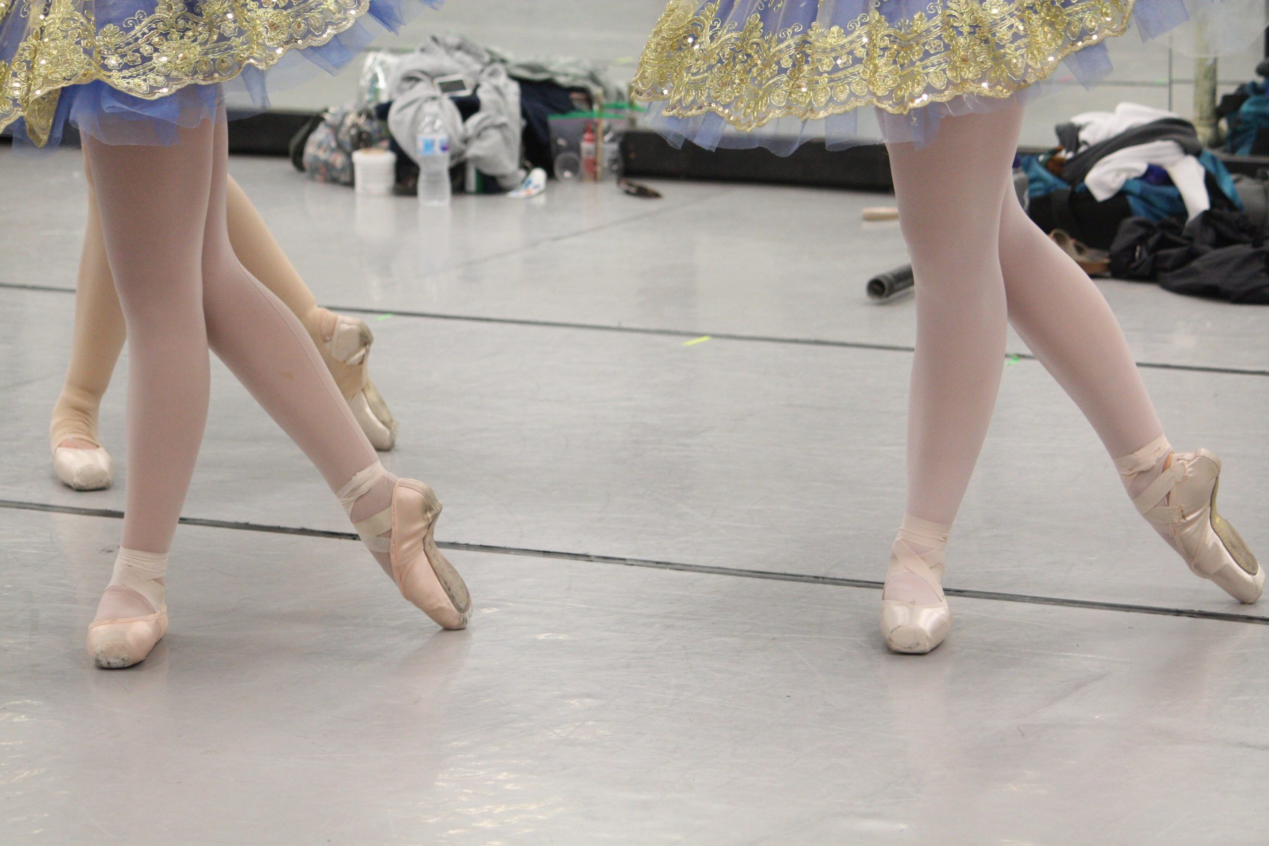 Close-up of ballet dancers’ feet in pointe shoes, standing in rehearsal on a marley floor.