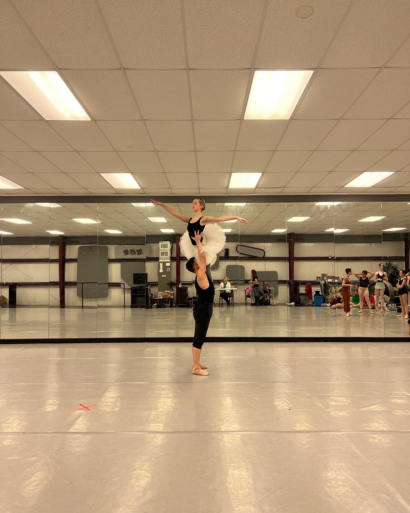 A young male ballet dancer in black lifts his female partner high above his head in a classical overhead lift during rehearsal. The studio’s mirrored wall reflects the strength, control, and artistry of the moment.