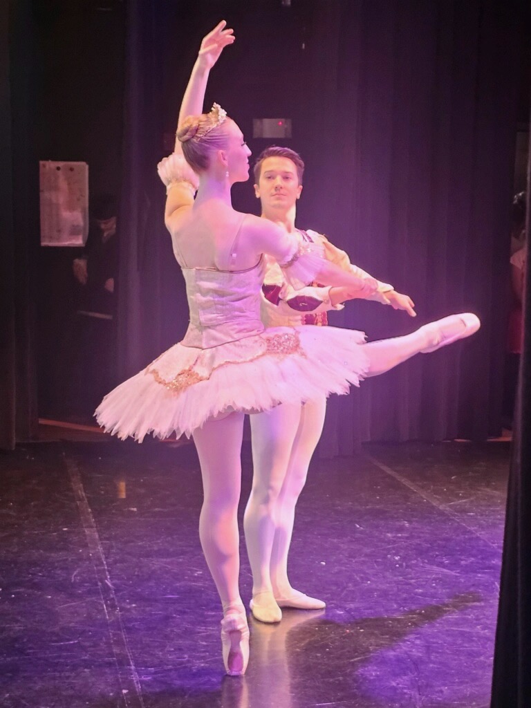 Two ballet dancers perform a pas de deux under pink and gold lighting. The male dancer, dressed as a prince, partners his ballerina in a sparkling white tutu, capturing the elegance and athleticism of male dancers in classical ballet.