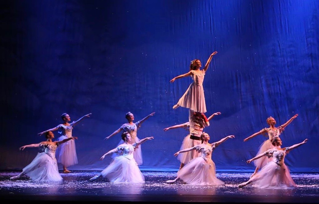 A group of ballerinas performs the snow scene from The Nutcracker under blue stage lighting. Dancers in white tutus surround the center where one dancer, lifted high above the others, extends her arms gracefully as snow falls around them.