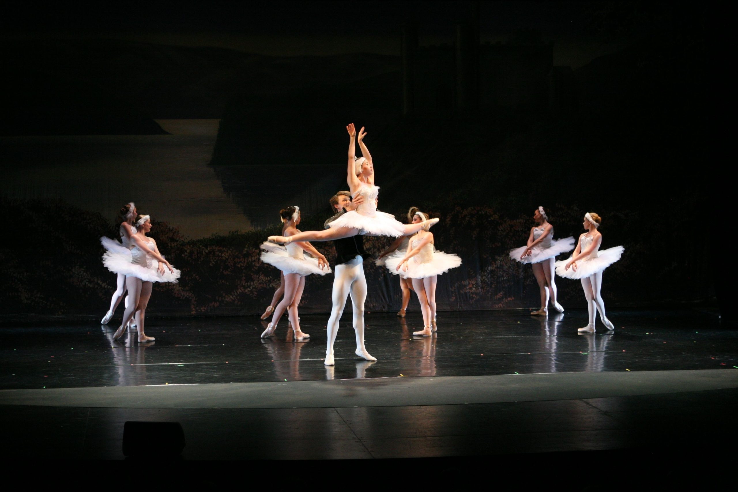 A male dancer in white tights lifts his partner in a grand overhead pose during a stage performance of Swan Lake. The stage is softly lit with a lake backdrop and a corps de ballet of swans surrounding them.