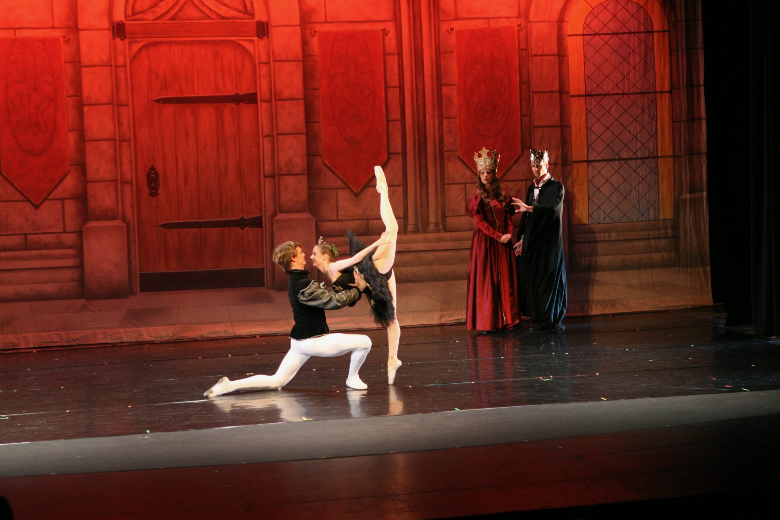 A male dancer in a period costume supports his partner in an elegant arabesque on stage, framed by a warm red castle backdrop. Two regal characters watch from the background, enhancing the storytelling drama of the scene.